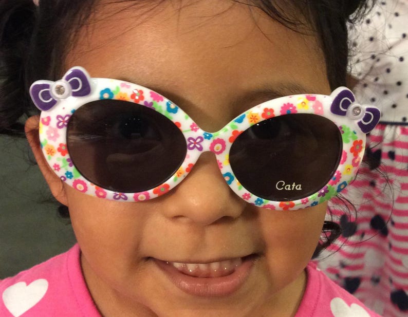 May include: A young girl wearing white sunglasses with a floral and colorful design. The sunglasses have a purple bow on each side and the text "Cata" is printed on the right lens.