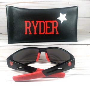 May include: Black sunglasses with red accents and a matching case. The case is black with the name "RYDER" in red and a white star. The sunglasses have a red emblem on the side.