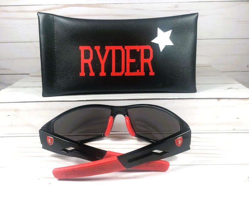 May include: Black sunglasses with red accents and a matching case. The case is black with the name "RYDER" in red and a white star. The sunglasses have a red emblem on the side.