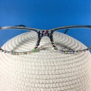 May include: Clear-framed eyeglasses adorned with colorful rhinestones. The rhinestones are in shades of pink, green, and blue, adding a touch of sparkle. The glasses are resting on a white, textured surface, with a blue background.