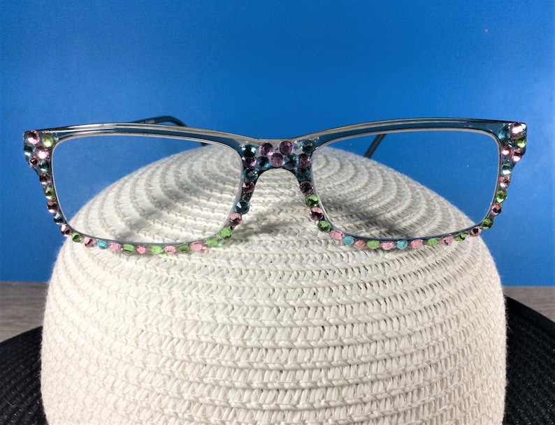 May include: Clear-framed eyeglasses adorned with colorful rhinestones. The rhinestones are in shades of pink, green, and blue, adding a touch of sparkle. The glasses are resting on a white, textured surface, with a blue background.