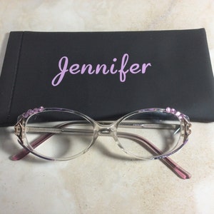 Puede incluir: Gafas transparentes con detalles rosas y pedrer&iacute;a decorativa en las monturas, frente a un estuche negro con el nombre "Jennifer" en escritura rosa.