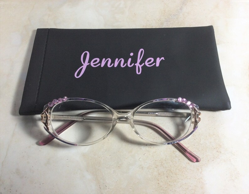 Puede incluir: Gafas transparentes con detalles rosas y pedrer&iacute;a decorativa en las monturas, frente a un estuche negro con el nombre "Jennifer" en escritura rosa.