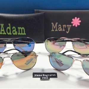 May include: Two pairs of aviator sunglasses with mirrored lenses. The sunglasses are in black cases with the names "Adam" and "Mary" printed on them. A sign reads "Name Engraved FREE".