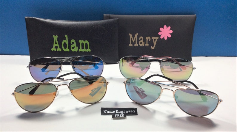 May include: Two pairs of aviator sunglasses with mirrored lenses. The sunglasses are in black cases with the names "Adam" and "Mary" printed on them. A sign reads "Name Engraved FREE".