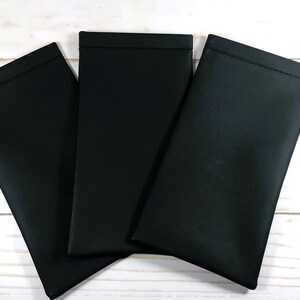 May include: Three black rectangular pouches are arranged on a white wooden surface. The pouches are made of a smooth, matte material and have a simple, minimalist design. The pouches are likely designed to hold glasses or other small items.