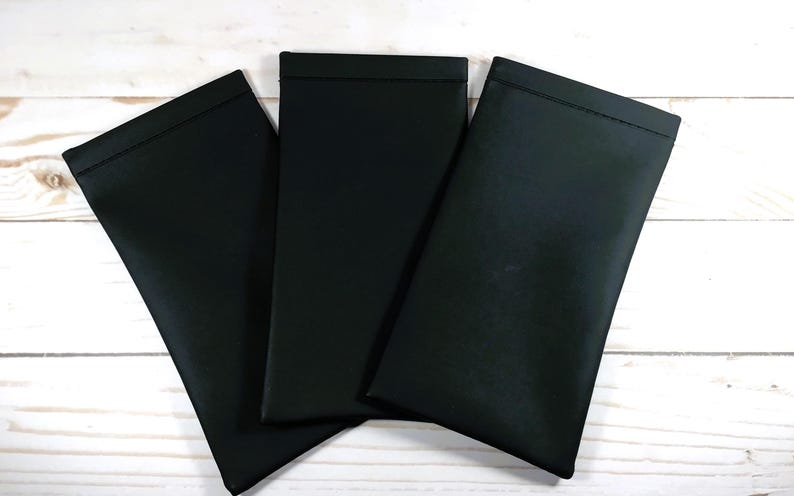 May include: Three black rectangular pouches are arranged on a white wooden surface. The pouches are made of a smooth, matte material and have a simple, minimalist design. The pouches are likely designed to hold glasses or other small items.