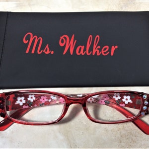 Puede incluir: Gafas rojas con estampado floral y detalles de pedrer&iacute;a, junto con un estuche negro. El estuche lleva la inscripci&oacute;n "Ms. Walker" en letras rojas.