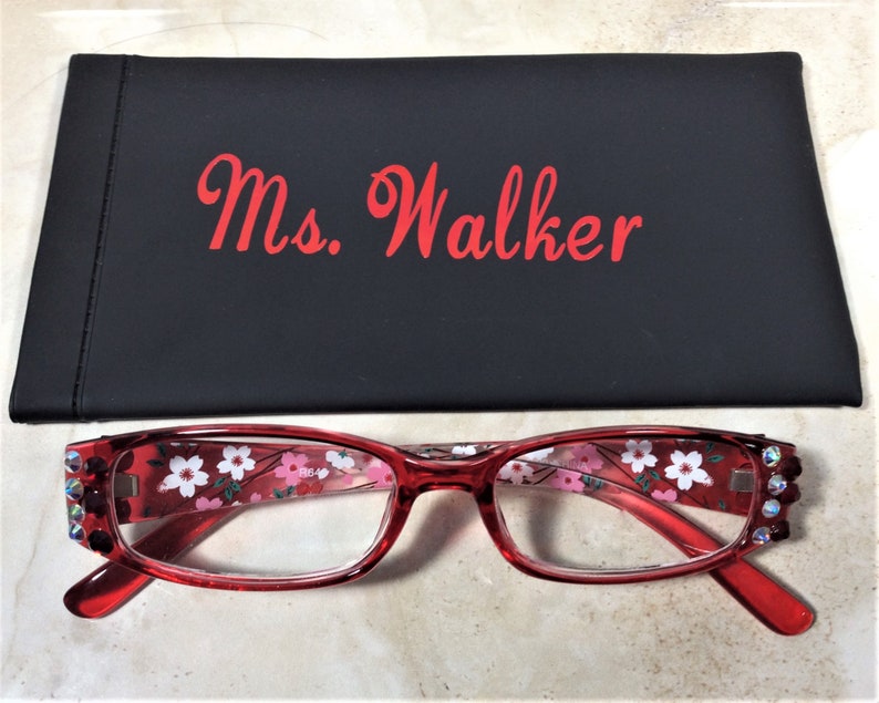 Puede incluir: Gafas rojas con estampado floral y detalles de pedrer&iacute;a, junto con un estuche negro. El estuche lleva la inscripci&oacute;n "Ms. Walker" en letras rojas.