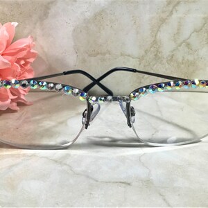 May include: Semi-rimless eyeglasses with a black frame and clear lenses. The top of the frame is adorned with iridescent rhinestones. Pink flowers are visible in the background. The glasses have a modern, stylish design.