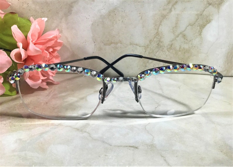 May include: Semi-rimless eyeglasses with a black frame and clear lenses. The top of the frame is adorned with iridescent rhinestones. Pink flowers are visible in the background. The glasses have a modern, stylish design.