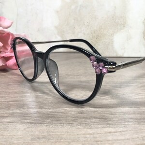 Puede incluir: Gafas de montura negra con forma redonda y varillas plateadas. Las monturas est&aacute;n adornadas con pedrer&iacute;a rosa en las sienes. Se encuentran sobre una superficie de madera, con un arreglo floral borroso en el fondo.