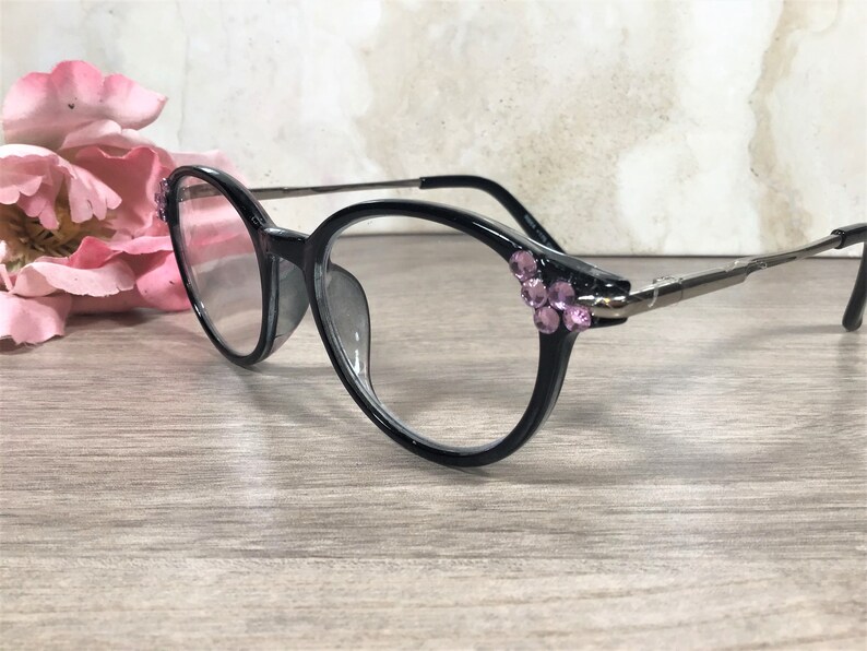 Puede incluir: Gafas de montura negra con forma redonda y varillas plateadas. Las monturas est&aacute;n adornadas con pedrer&iacute;a rosa en las sienes. Se encuentran sobre una superficie de madera, con un arreglo floral borroso en el fondo.