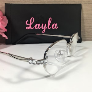 K&ouml;nnte beinhalten: Silberfarbene Brille mit klaren Gl&auml;sern und Strasssteinen entlang des Rahmens. Die Brille wird mit einem schwarzen Etui mit dem Namen "Layla" in rosa Schrift gezeigt. Rosa Blumen sind im Hintergrund.