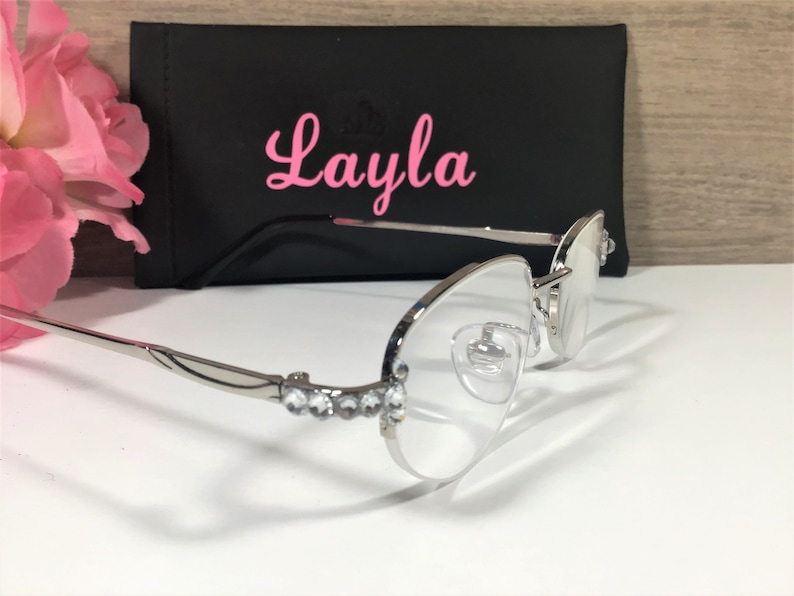 K&ouml;nnte beinhalten: Silberfarbene Brille mit klaren Gl&auml;sern und Strasssteinen entlang des Rahmens. Die Brille wird mit einem schwarzen Etui mit dem Namen "Layla" in rosa Schrift gezeigt. Rosa Blumen sind im Hintergrund.