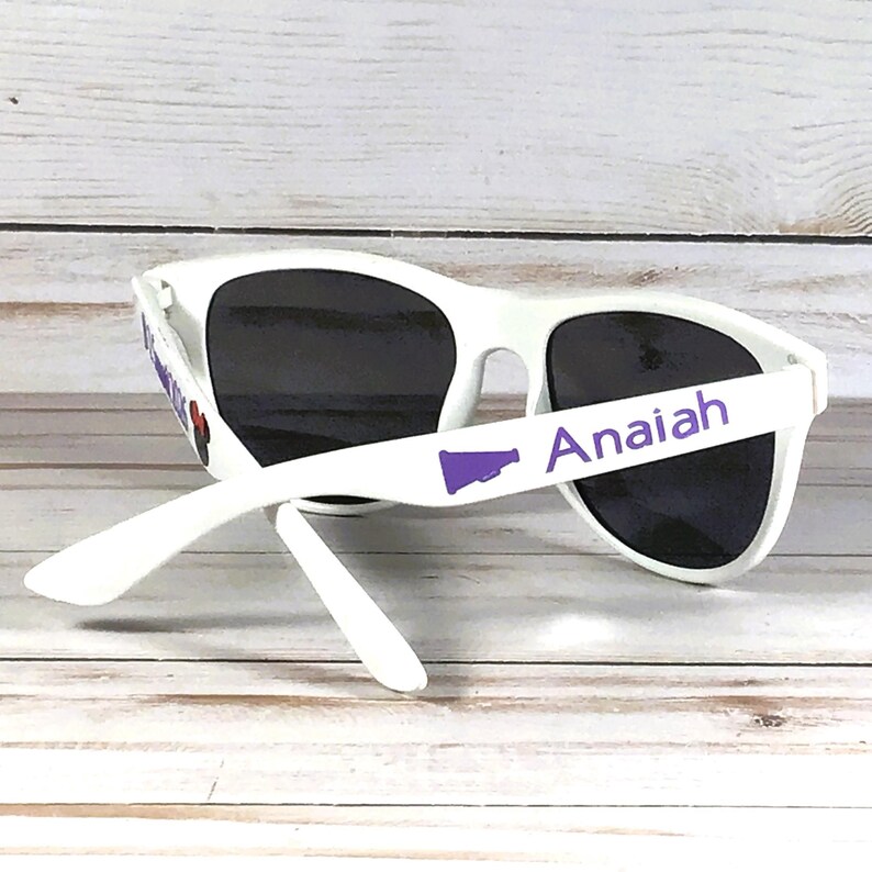May include: White sunglasses with black lenses. The right arm of the sunglasses has the name "Anaiah" printed in purple, along with a purple megaphone graphic. The left arm has a purple design.