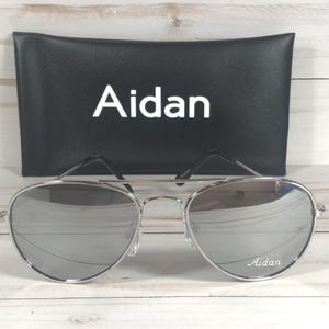May include: Silver aviator sunglasses with a thin metal frame and reflective lenses. The right lens features the white text Aidan. Behind the sunglasses sits a black rectangular protective case with the white text Aidan printed in the center.