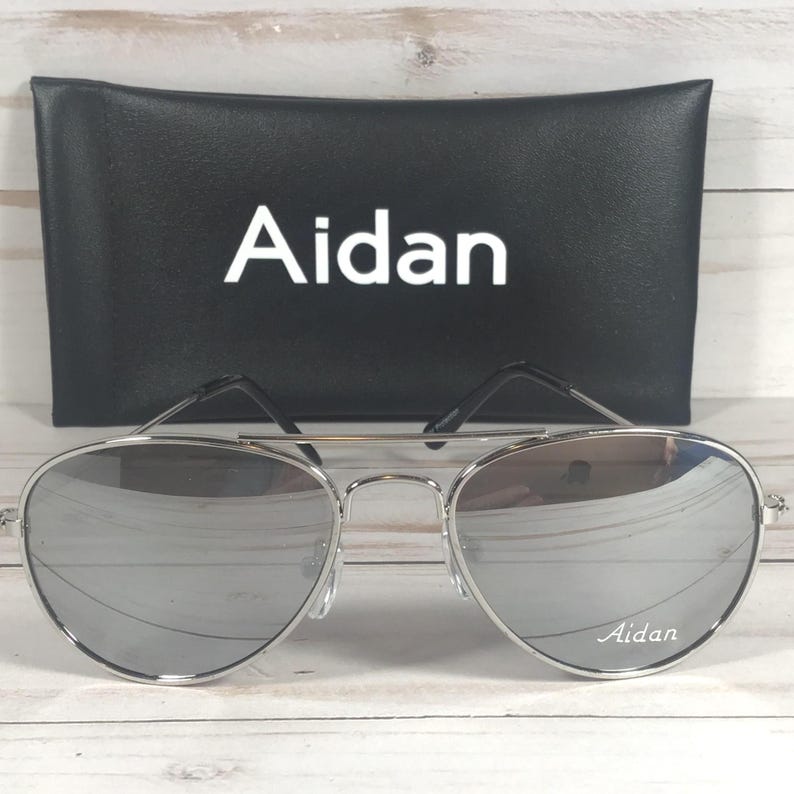 May include: Silver aviator sunglasses with a thin metal frame and reflective lenses. The right lens features the white text Aidan. Behind the sunglasses sits a black rectangular protective case with the white text Aidan printed in the center.