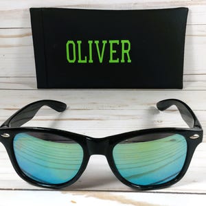 Pu&ograve; includere: Occhiali da sole neri con lenti a specchio verdi e una custodia nera con il nome "OLIVER" stampato in verde.