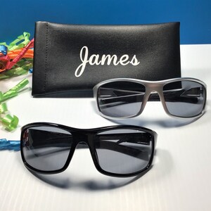 K&ouml;nnte beinhalten: Zwei Sonnenbrillen, eine schwarze und eine silberne, mit einem schwarzen Sonnenbrillenetui, auf dem "James" steht.