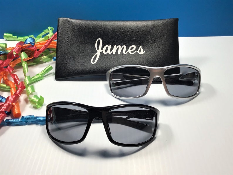 K&ouml;nnte beinhalten: Zwei Sonnenbrillen, eine schwarze und eine silberne, mit einem schwarzen Sonnenbrillenetui, auf dem "James" steht.