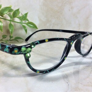 K&ouml;nnte beinhalten: Schwarze Brille mit floralem Muster in T&uuml;rkis und Gelb. Die Fassungen sind mit gr&uuml;nen Strasssteinen verziert. Die Brille hat klare Gl&auml;ser und schwarze B&uuml;gel.