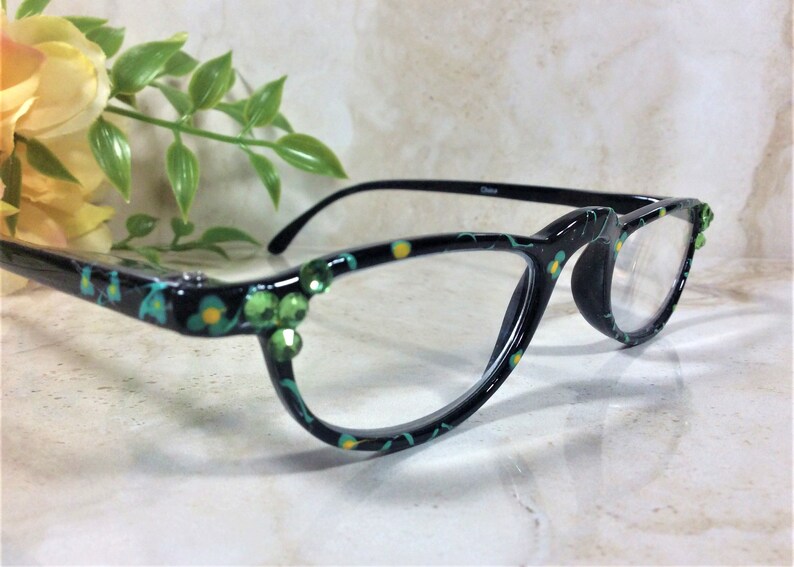 K&ouml;nnte beinhalten: Schwarze Brille mit floralem Muster in T&uuml;rkis und Gelb. Die Fassungen sind mit gr&uuml;nen Strasssteinen verziert. Die Brille hat klare Gl&auml;ser und schwarze B&uuml;gel.