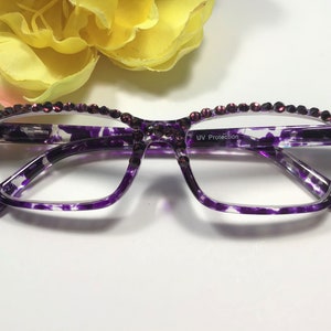 May include: Purple-framed reading glasses with a decorative rhinestone border. The frames have a marbled purple and white pattern. The glasses have UV protection.