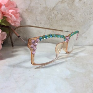 May include: A pair of eyeglasses with a light orange frame and clear lenses. The frame is adorned with pink, blue, and green rhinestones. The glasses have a gold-colored metal arm and are set against a marble background.