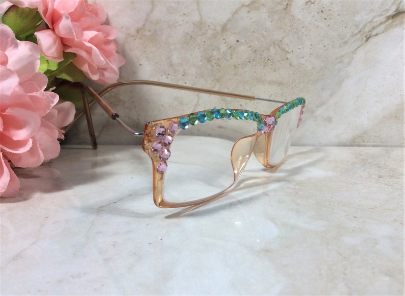May include: A pair of eyeglasses with a light orange frame and clear lenses. The frame is adorned with pink, blue, and green rhinestones. The glasses have a gold-colored metal arm and are set against a marble background.