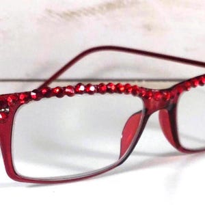 May include: Red-framed eyeglasses with clear lenses. The top of the frames are embellished with small, sparkling red rhinestones. The temples are thin and extend to the ear pieces. The glasses are set against a white background.