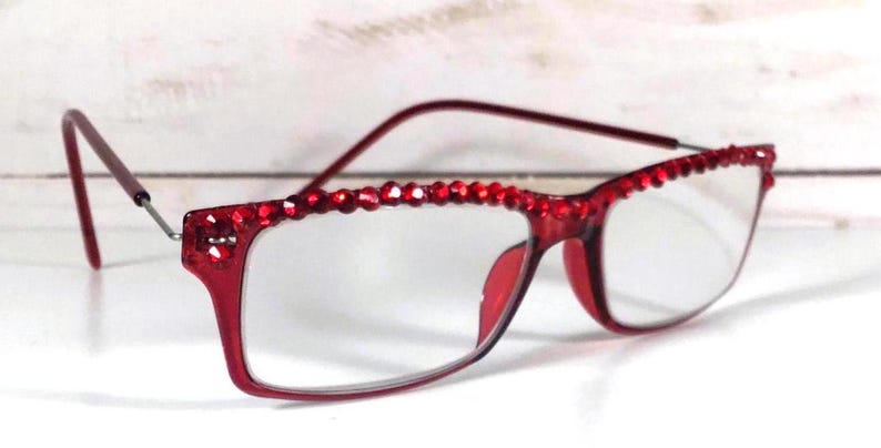 May include: Red-framed eyeglasses with clear lenses. The top of the frames are embellished with small, sparkling red rhinestones. The temples are thin and extend to the ear pieces. The glasses are set against a white background.