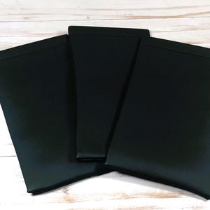 May include: Three black fabric items, possibly cases or covers, are arranged on a light wood-grain surface. The items are rectangular with a folded top edge, and they are overlapping slightly. The fabric has a matte finish.