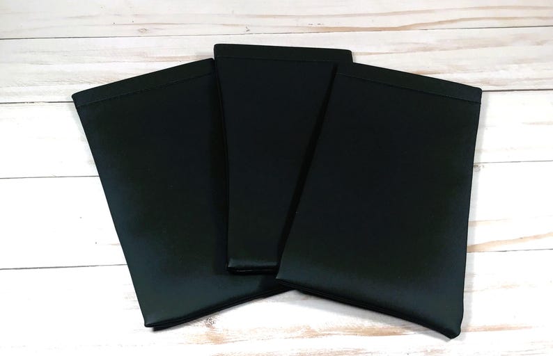 May include: Three black fabric items, possibly cases or covers, are arranged on a light wood-grain surface. The items are rectangular with a folded top edge, and they are overlapping slightly. The fabric has a matte finish.