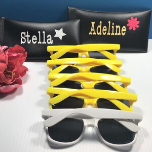 May include: A stack of four pairs of bright yellow sunglasses and one pair of white sunglasses, positioned in front of two black rectangular cases. One case features the name Stella with a star, and the other features Adeline with a flower.