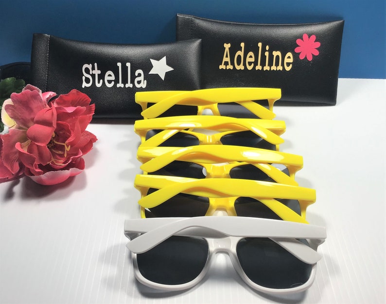May include: A stack of four pairs of bright yellow sunglasses and one pair of white sunglasses, positioned in front of two black rectangular cases. One case features the name Stella with a star, and the other features Adeline with a flower.