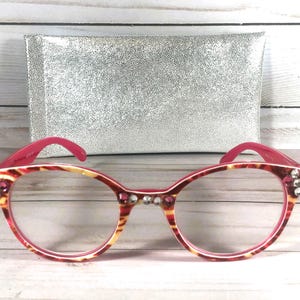 May include: Cat-eye glasses featuring a pink and yellow tortoiseshell pattern on the frames. The front of the frames is embellished with clear and pink rhinestones at the temples and bridge. A rectangular silver glitter case is positioned behind.