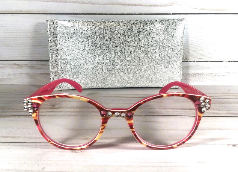 May include: Cat-eye glasses featuring a pink and yellow tortoiseshell pattern on the frames. The front of the frames is embellished with clear and pink rhinestones at the temples and bridge. A rectangular silver glitter case is positioned behind.