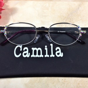 May include: Oval silver-rimmed spectacles with clear lenses, embellished with iridescent rhinestones at the temples. The glasses sit on a black case with the name "Camila" in white. A pink rose is in the background.