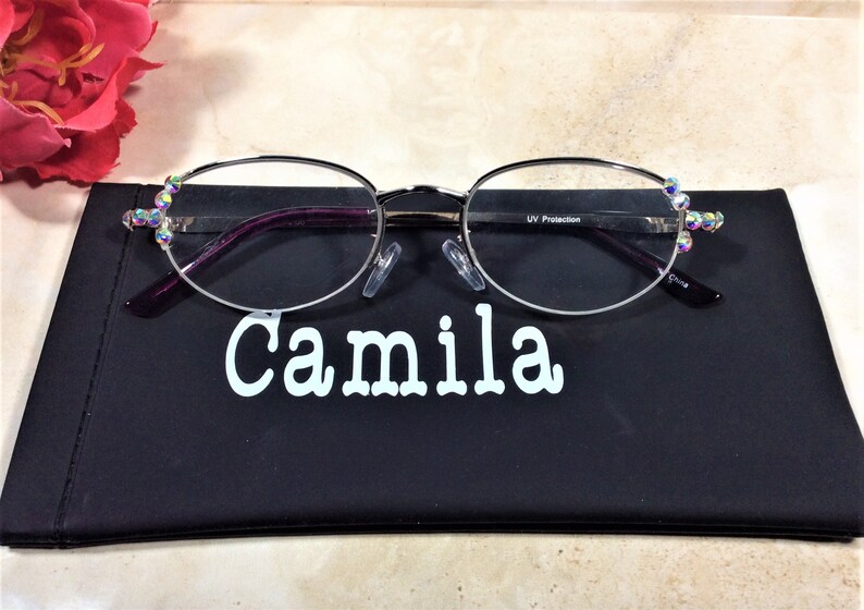 May include: Oval silver-rimmed spectacles with clear lenses, embellished with iridescent rhinestones at the temples. The glasses sit on a black case with the name "Camila" in white. A pink rose is in the background.