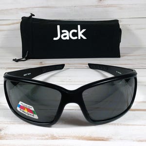 Puede incluir: Gafas de sol envolventes negras con lentes oscuras y una peque&ntilde;a pegatina rectangular en la lente izquierda que dice 100% Ultra Violet Protection Polarized. Detr&aacute;s de las gafas hay una bolsa de tela negra con el texto blanco Jack impreso.