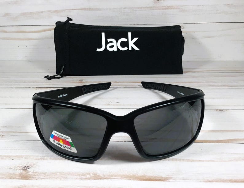 Puede incluir: Gafas de sol envolventes negras con lentes oscuras y una peque&ntilde;a pegatina rectangular en la lente izquierda que dice 100% Ultra Violet Protection Polarized. Detr&aacute;s de las gafas hay una bolsa de tela negra con el texto blanco Jack impreso.