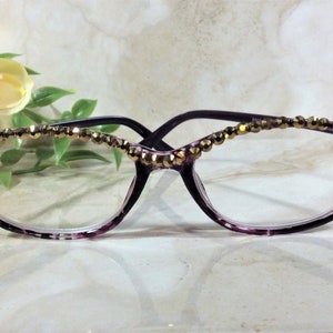 May include: Purple spectacles featuring a translucent frame with a tortoiseshell pattern. The upper rim of the frame is embellished with a single row of small, faceted, metallic gold-toned beads that catch the light across the top edge.