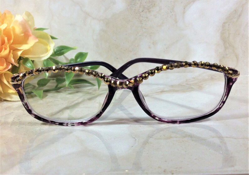 May include: Purple spectacles featuring a translucent frame with a tortoiseshell pattern. The upper rim of the frame is embellished with a single row of small, faceted, metallic gold-toned beads that catch the light across the top edge.