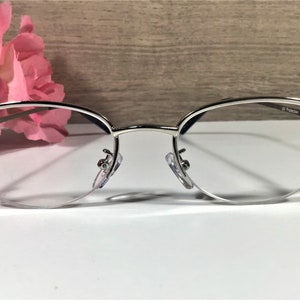 K&ouml;nnte beinhalten: Brille mit silbernem Rahmen und klaren Gl&auml;sern. Die Fassungen haben eine zarte, ovale Form und sind mit dekorativen Akzenten an den B&uuml;geln verziert. Die Gl&auml;ser haben einen UV-Schutz. Die Brille liegt auf einer wei&szlig;en Oberfl&auml;che.