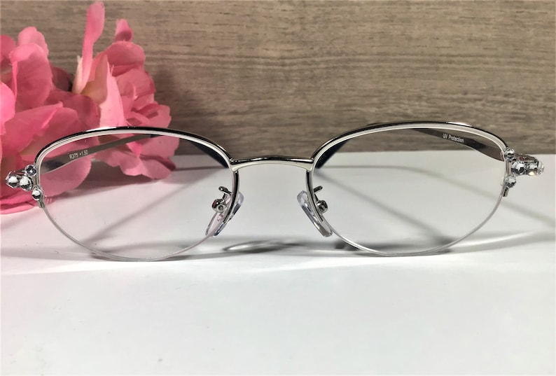 K&ouml;nnte beinhalten: Brille mit silbernem Rahmen und klaren Gl&auml;sern. Die Fassungen haben eine zarte, ovale Form und sind mit dekorativen Akzenten an den B&uuml;geln verziert. Die Gl&auml;ser haben einen UV-Schutz. Die Brille liegt auf einer wei&szlig;en Oberfl&auml;che.