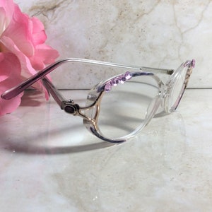 Puede incluir: Gafas transparentes con un dise&ntilde;o decorativo. Las monturas son transparentes con detalles dorados y adornadas con peque&ntilde;os brillantes rosas. Las patillas son de color lila claro. Una flor rosa est&aacute; en el fondo.