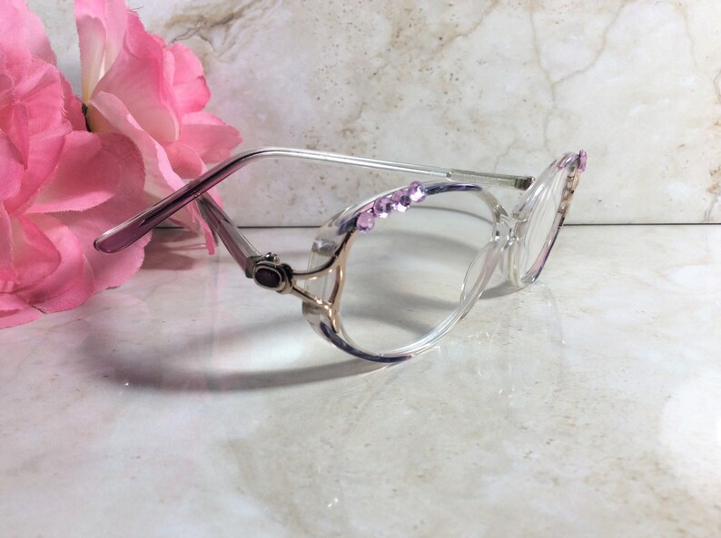 Puede incluir: Gafas transparentes con un dise&ntilde;o decorativo. Las monturas son transparentes con detalles dorados y adornadas con peque&ntilde;os brillantes rosas. Las patillas son de color lila claro. Una flor rosa est&aacute; en el fondo.