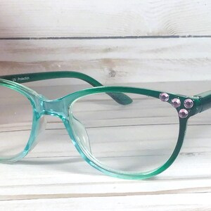 May include: Translucent teal green eyeglasses featuring a gradient frame design. The outer corners of the frames are embellished with small, round, light purple rhinestones. The inner temple arm includes the printed text UV Protection.