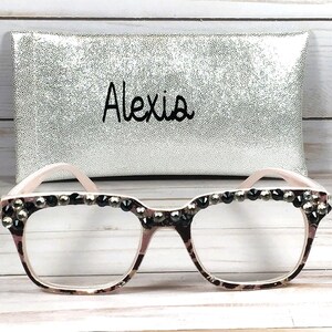May include: Eyeglasses with a patterned frame and embellished with dark rhinestones. The glasses are paired with a silver pouch with the name "Alexia" in black script.